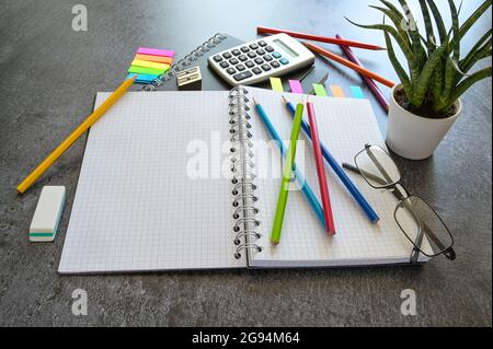 Büro- oder Schreibtisch mit offenem Spiralheftchen, Buntstiften, Taschenrechner und Schreibwaren auf einem dunkelgrauen Tisch, Kopierfläche, ausgewähltem Fokus Stockfoto