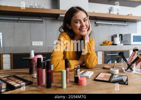 Lächelnde junge Brünette Frau macht Make-up, während sie in der Küche zu Hause sitzt Stockfoto