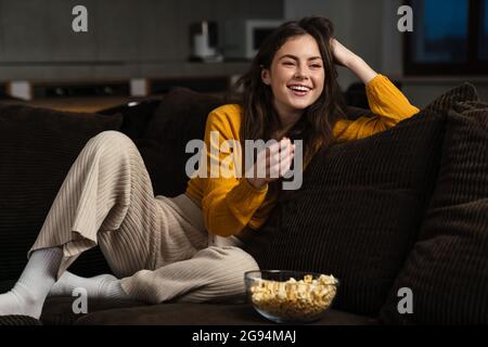 Lächelnde junge Brünette Frau, die Popcorn isst, während sie zu Hause auf einer Couch sitzt Stockfoto