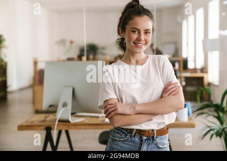 Nahaufnahme einer lächelnden jungen Brünette, die lässig gekleidet im Büro steht, die Arme gefaltet Stockfoto