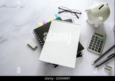 Schreibtisch mit leerem Notizbuch, Sparschwein, Taschenrechner und stationärem Zubehör auf Marmoroberfläche, Kopierraum, ausgewählter Fokus, enge Tiefe des Fie Stockfoto