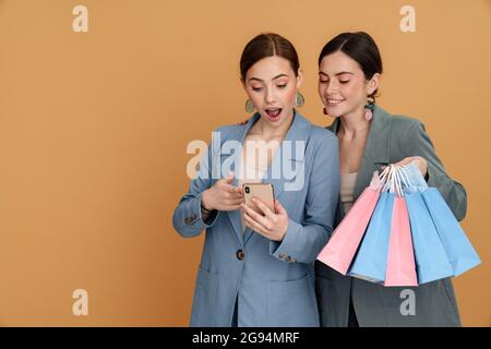 Zwei junge Frauen, die Einkaufstaschen in der Hand halten und auf gelbem Hintergrund ein isoliertes Mobiltelefon benutzen Stockfoto