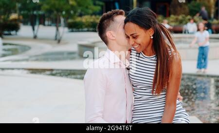 Nahaufnahme des jungen Mannes, der etwas im Ohr der Freundin flüstert, das draußen auf einer Bank sitzt. Sie lächeln, umarmen und reden miteinander. Stockfoto