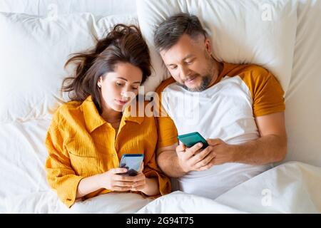 Nahaufnahme eines Paares mittleren Alters, das mit Smartphones im Bett liegt. Konzept moderner Technologien im Alltag. Stockfoto