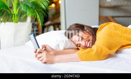 Glückliche Frau mit Telefon im Bett. Sie lacht glücklich. Modernes Technologiekonzept. Stockfoto