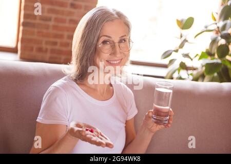 Portrait einer attraktiven, fröhlichen, grauhaarigen Frau, die Wasser trinkt und Pillen zu Hause im modernen Backsteininterieur nimmt Stockfoto