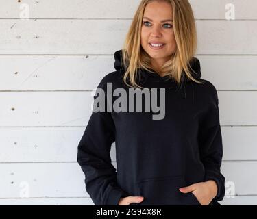 Eine lächelnde Blondine steht in einem schwarzen Sweatshirt auf einem Hintergrund aus weißen Brettern vor der Kamera. Auf dem Kleidungsstück befindet sich ein leerer Platz für ein Design, ein Logo oder eine Beschriftung. Nachbau für Kleidung. Stockfoto