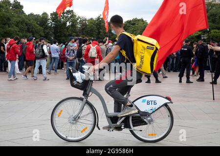 Moskau, Russland. 24. Juli 2021 vor dem Hintergrund einer kommunistischen Kundgebung, Russland, liefert der Kurier des Lebensmittellieferdienstes Yandex.EDA einen Auftrag an einen Kunden in der Tverskaya-Straße im Zentrum von Moskau Stockfoto