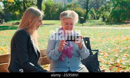 Ältere Großmutter, die ihre Enkelin auf dem Smartphone zeigt. Sie sitzen auf der Bank und reden im Herbstpark. Stockfoto
