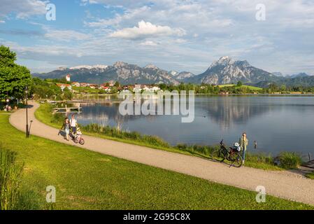 Am Ufer des Hopfensees wandern die Menschen vor den Bergen der Ammergauer Alpen in der Nachmittagssonne, Allgäu, Bayern, Deutschland Stockfoto