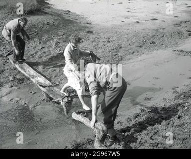 1950s, historisch, Schuhe aus, eine Dame in barfuß, als sie einen flachen Bach überquert, unterstützt von ihren Geherkollegen, einer stehend auf einem Holzbrett, der andere einen hölzernen Ast haltend und ihr seine ausgestreckte Hand anbietet, während sie hinüber geht, auf einem sandigen Gebiet in der Nähe von Beachy Head, Eastbourne, East Sussex, England, VEREINIGTES KÖNIGREICH. Stockfoto