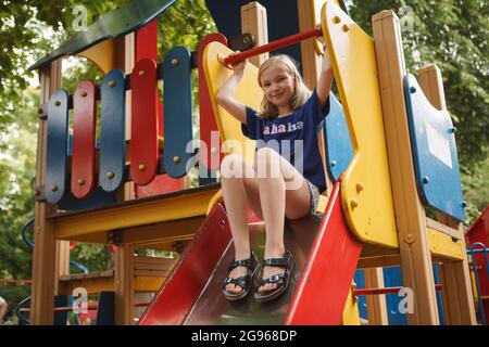 Low-Angle-Aufnahme eines fröhlichen Mädchens, das auf der Rutsche auf dem Spielplatz sitzt und lächelt zur Kamera Stockfoto