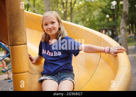 Charming glückliches Mädchen gleiten auf Spielplatz Rutsche, lächelnd, um die Kamera Stockfoto