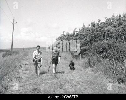 1950er, historisch, sommerlich und zwei männliche Wanderer mit einem Hund, die entlang eines grasbewachsenen Pfades auf den South Downs, East Sussex, England, Großbritannien, wandern. Die hügeligen South Downs sind bekannt für Beachy Head, die höchste kalkförmige Meeresklippe Großbritanniens, und sind ein berühmter Ort für Spaziergänge. Ein Gebiet von außergewöhnlicher natürlicher Schönheit, im Jahr 1929, ein Akt des Parlaments ermöglichte es, das Land zu kaufen, um seine Nutzung für zukünftige Generationen zu sichern. Stockfoto