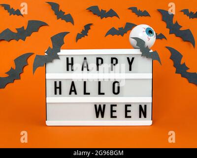 Leuchtkasten mit Happy Halloween Phrase mit Fledermäusen und Augapfel-Dekoration auf orangefarbenem Hintergrund. Stockfoto