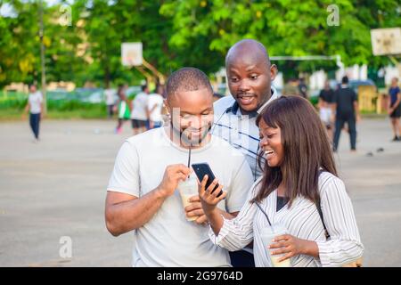 Fröhliche Gruppe afrikanischer Freunde, bestehend aus zwei Jungs und einer Dame, die freudig ein Smartphone in einem Park im Freien anschaut, während sie jubelt Stockfoto