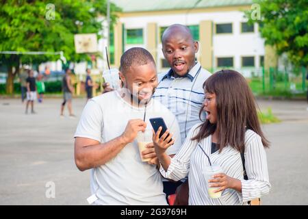 Fröhliche Gruppe afrikanischer Freunde, bestehend aus zwei Jungs und einer Dame, die freudig ein Smartphone in einem Park im Freien anschaut, während sie jubelt Stockfoto