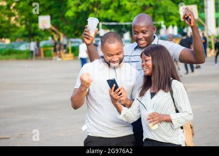 Fröhliche Gruppe afrikanischer Freunde, bestehend aus zwei Jungs und einer Dame, die freudig ein Smartphone in einem Park im Freien anschaut, während sie jubelt Stockfoto