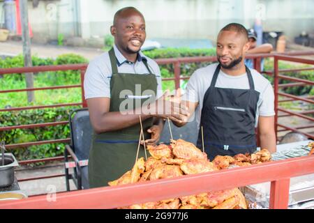 Zwei männliche afrikanische Barbeque- und Hühnerverkäufer lächeln und begrüßen Kunden, um ihr Geschäft zu bevormunden Stockfoto