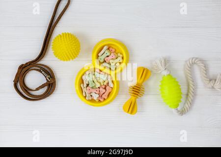 Gelbe Schale mit Snacks, Ball, Leine, ein Knochenspielzeug für Hunde auf weißem Holztisch. Set von Zubehör für Haustiere auf Holz. Nahaufnahme Stockfoto