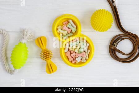 Gelbe Schale mit Snacks, Ball, Leine, ein Knochenspielzeug für Hunde auf weißem Holztisch. Set von Zubehör für Haustiere auf Holz. Nahaufnahme Stockfoto