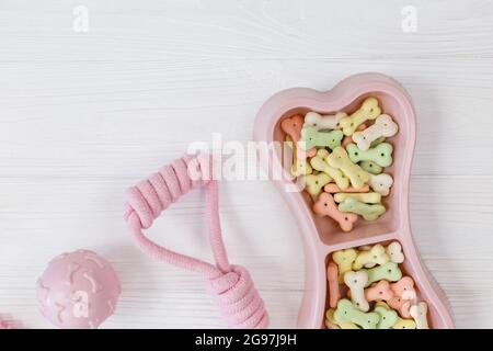 Rosa Schale mit Snacks, Ball, Leine, ein Knochenspielzeug für Hunde auf einem weißen Holztisch. Set von Zubehör für Haustiere auf Holz. Nahaufnahme Stockfoto