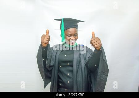 Stock-Foto einer afrikanischen Graduiertenin oder einer Absolventin in ihrem schwarzen Abschlussexamen-Outfit und freudig mit Daumen nach oben Geste Stockfoto
