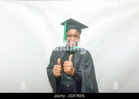 Stock-Foto einer afrikanischen Graduiertenin oder einer Absolventin in ihrem schwarzen Abschlussexamen-Outfit und freudig mit Daumen nach oben Geste Stockfoto