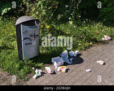 Mülltonne im Parkgelände in Koblenz mit Verpackungsmüll, der am sonnigen Sommertag mit Gras und Büschen herumliegt. Stockfoto