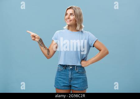 Studio Aufnahme von fasziniert charmante junge europäische Frau mit gebräunt Haut und blonde Haare halten Hand an der Taille in zuversichtlich Pose suchen und zeigen Stockfoto