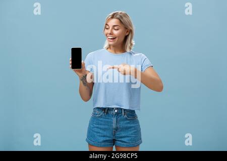 Portrait der entzückten gut aussehenden europäischen Frau mit blonden Haarschnitt zeigt Smartphone-Bildschirm mit Erstaunen und Freude zeigt auf Gerät während Stockfoto