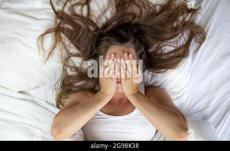 Junge Frau, die im Bett liegt, leidet, müde Frau, die das Gesicht mit den Händen bedeckt, kann nicht schlafen, fühlt sich auf weißen Laken im Schlafzimmer erschöpft Stockfoto