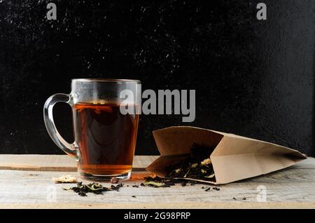 Ein Glas mit duftendem, gebrühtem Blumentee auf einem Holztisch, mit einem Teeblätter in einem Papierbeutel. Selektiver Fokus. Stockfoto