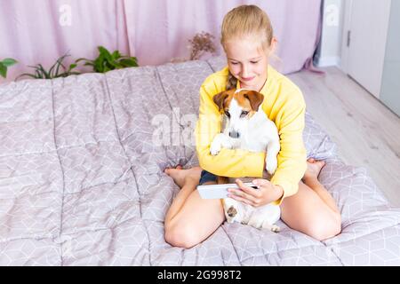 Ein Teenager-Mädchen mit einem Mobiltelefon, umarmt einen Hund Jack Russell Terrier, kommuniziert in sozialen Netzwerken oder via Zoom mit Freunden oder Familie, macht eine Stockfoto