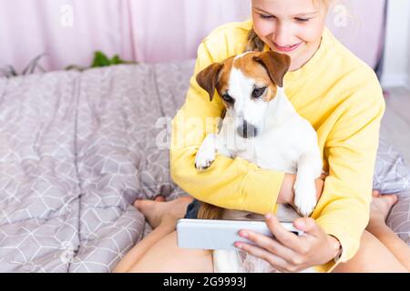 Ein lächelndes Mädchen mit einem Handy in der Hand, zusammen mit ihrem Hund Jack Russell Terrier, kommuniziert in sozialen Medien oder via Zoom, macht ein Selfie Stockfoto