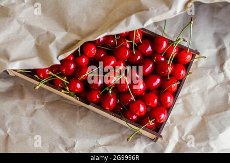 Cropped Box, Crape von dunkelroten süßen Kirschen mit Schwanz auf Handwerk zerknittert Papier Hintergrund. Sommerfrüchte und Beeren. Ernte- und Erntekonzept Stockfoto