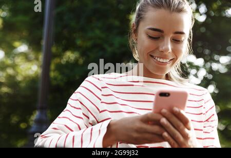 Porträt eines lächelnden blonden Mädchens, das auf dem Handy chattet, auf der Bank im Park sitzt und das Smartphone benutzt Stockfoto