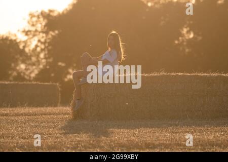 Latina Teenager-Mädchen mit langen Haaren und weißem T-Shirt auf einem Strohballen in einem geernteten Getreidefeld sitzen. Sonne leuchtet während des Sonnenuntergangs, warmes Bild. Stockfoto