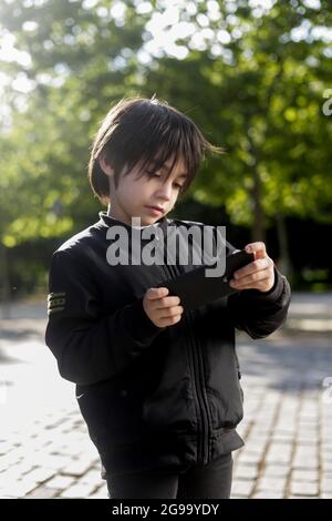 8-jähriger hispanischer Junge, der mit einem Mobiltelefon im Park spielt Stockfoto