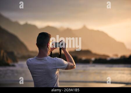 Rückansicht des Menschen beim Fotografieren von Landschaft mit Klippe. Fotograf am Strand bei stimmungsvollen Sonnenuntergängen. Teneriffa, Kanarische Inseln, Spanien. Stockfoto