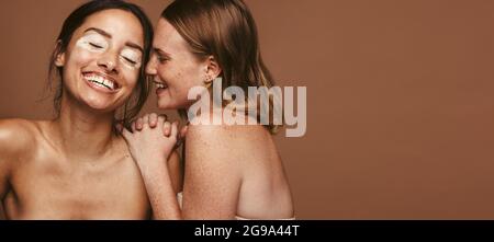 Porträt zweier junger Frauen mit verschiedenster Haut, die auf braunem Hintergrund zusammenstehen. Junge Frauen mit Vitiligo und Sommersprossen in fröhlicher Stimmung represe Stockfoto