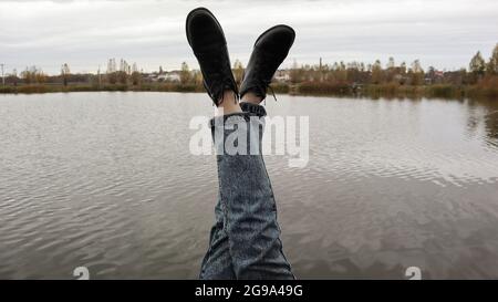 Ein Mädchen in blauen Jeans sitzt am See. Eine junge Frau ruht am Fluss und sitzt am Rande einer Holzbrücke. Zwei Beine, die sich an einem See entspannen Stockfoto