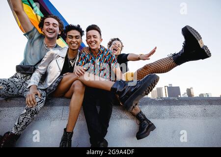 Vier LGBTQ-Menschen feiern Stolz, während sie zusammen sitzen. Vier Freunde lächeln fröhlich, während sie die Regenbogenflagge heben. Gruppe junger Queer Stockfoto