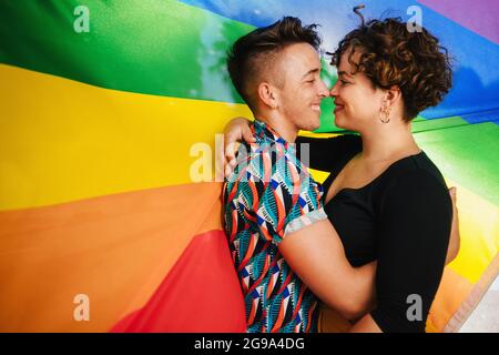 Romantisches queeres Paar, das sich gegen eine Regenbogenflagge festmacht. Das junge LGBTQ-Paar umarmt sich gegenseitig und berührt ihre Nasen. Zwei nicht konform Stockfoto