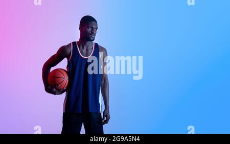 Cooler schwarzer Basketballspieler in Sportswear, der den Ball hält und im Neonlicht-Bannerdesign auf leeren Raum schaut Stockfoto