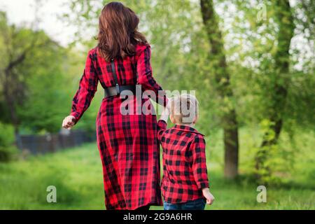 Mutter auf Spaziergang mit ihrem Sohn im Park. Frau und Kind in der Natur. Rückansicht. Familienlook. Rustikal. Der Elternteil hält die Hand des Kindes. Internationales Konzept für Familientag Stockfoto