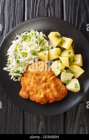 Polnische Küche Kotlet Schabowy mit gekochten Kartoffeln und Kohlsalat in Nahaufnahme auf einem Teller auf schwarzem Hintergrund. Vertikale Draufsicht von oben Stockfoto