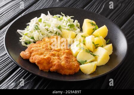 Gebratenes Schweinekotelett paniert mit Kartoffeln und Kohlsalat in Nahaufnahme auf einem Teller auf schwarzem Holzhintergrund. Horizontal Stockfoto