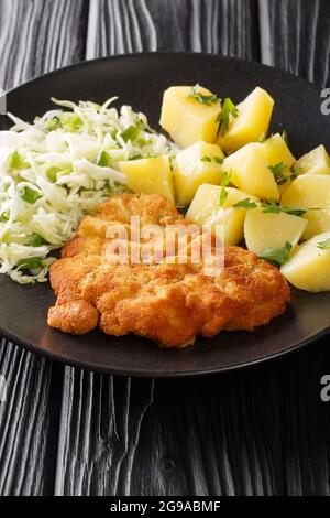 Kotlet Schabowy Polnischer panierter Schweineball mit gekochten Kartoffeln und Kohlsalat in Nahaufnahme auf einem Teller auf einem schwarzen Tisch. Vertikal Stockfoto