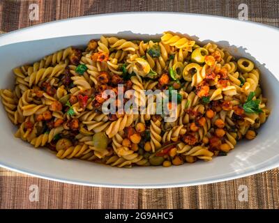 Nahaufnahme von Nudelgerichten: Fusilli mit Kichererbsen, Kapern, Oliven und Tomaten in weißer Auflaufform auf dem Freilufttisch bei Sonnenschein mit Weinflasche serviert Stockfoto